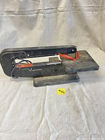 Side view of Dremel 571-5 Moto-Shop scroll saw showing black metal frame, blade area with orange tensioner, flat work surface, and power cord.