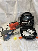 Three electric sanders displayed on cloth background including Ryobi RS290, Black & Decker RO600 SandStorm with case and manual, Black & Decker Mouse detail sander with sanding pads and case.