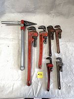 Photo of six Ridgid pipe wrenches of varying sizes, mostly red with metal jaws, arranged on a cloth background, alongside a metal pipe reamer with T-handle and spiral cutting head.
