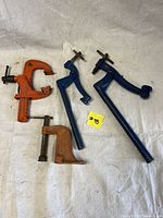 Four woodworking clamps including two orange C-clamps and two blue hold-down clamps arranged on a cloth surface.
