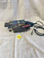 Side angle showing blue Ryobi belt sander with dust bag and power cord coiled beside it, placed on a white textured cloth.