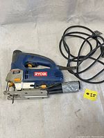 Side view of Ryobi JS550L jig saw showing blue plastic housing with Ryobi logo, orange button, metal base plate, and cord with dust collection tube duct taped on.