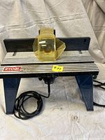 Front view of the RYOBI RT102 router table with power cord coiled in front and the yellow safety guard on the table surface.