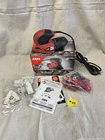 SKIL Octo 7302 sander kit with sander, box, manuals, and red clips all displayed on beige cloth background.