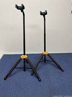 Two black Hercules guitar stands upright on blue padded surface, showing legs fully extended and top U-shaped supports.
