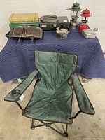 Overall view of the camping equipment showing stoves, lanterns, kettle, hibachi, camp chair, and cooler box.