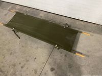Full view of the collapsible stretcher laid flat on concrete floor showing olive green canvas bed and wooden carrying handles.