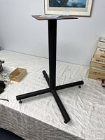 Full view of black metal table base showing the square mounting plate, central column, and four extending legs on a white patterned surface with background items.