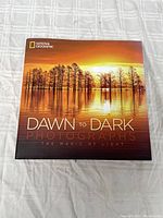 Front cover of the National Geographic Dawn to Dark photograph book showing an orange sunset reflection with silhouetted trees.
