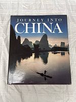 Front cover of the National Geographic Journey Into China photographic hardcover book showing karst mountain scenery and a person on a boat at sunset/sunrise.