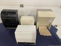 Front view of 4 wall-mount paper towel and sheet dispensers on a blue padded surface, showing one black dispenser and three white/cream dispensers.