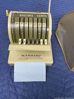 Front view of the antique Paymaster cheque writer showing rotary dials for dollars and cents and currency symbols with a blue paper inserted.