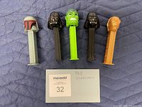 Front view of five vintage PEZ candy dispensers lined up on quilted fabric, with MaxSold card labeled 32 in front of them