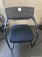 Front view of a single black stacking chair showing plastic perforated back, cloth seat cushion, and steel frame.