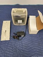 Front view of the Simplex 6200 time clock unit with key inserted, adjacent to the metal wall mounting rack, AC power adapter, and box of time cards opened to show cards.