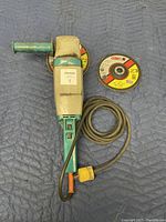 Full view of Wolf Sapphire grinder/sander laid flat with power cord coiled, green handle visible, two 7-inch CGW branded abrasive discs partially underneath and beside tool.