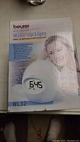 Front view of Beurer WL32 Wake-Up Light box showing product image, digital clock display, and highlights of natural sunlight simulation for waking up.