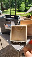 Photo showing black collapsible rolling crate with retractable handle and two wooden boxes (one large box and one small box with rope handles) on a table in outdoor setting.
