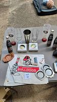 Full lot table showing Georgia Bulldogs themed glasses, Coca-Cola bottles, lighters, towel with mascot, bar tools and collectibles.