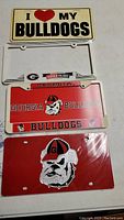 Four Georgia Bulldogs license plates and one frame laid out on a white surface. Plates show various logos and text. One plate is sealed in plastic.