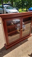 Wooden hutch with two large glass doors and brass knobs, photographed outside with reflections of vehicles.