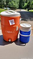 Two water coolers side by side; large orange Igloo cooler with white lid and spigot, smaller blue and white Coleman cooler.