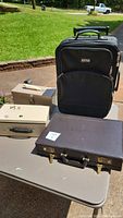 Four items displayed outdoors: two metal lockboxes, a hard briefcase, and a wheeled luggage.