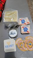 Photo of vintage beer coasters and napkins including stacked Pabst Blue Ribbon coasters, Newcastle Brown Ale coasters, Merit napkins and sealed pack of Meeot napkins