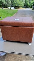 Brown faux leather ottoman with hinged top and silver metal legs placed on a table outdoors with a road and trees in background