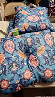 Three blue outdoor throw pillows with multicolored floral designs stacked with some tags visible showing they are new.