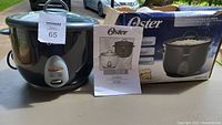 Front view of Oster rice cooker with box and manual in background on outdoor table surface.