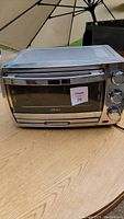 Full view of Oster toaster oven on wooden surface showing the stainless steel exterior and glass door with the MaxSold lot sticker.