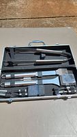 Full view of grilling set tool open inside metal carrying case showing stainless steel utensils with textured metal handles securely placed in foam insert