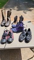Full lot view showing five pairs of shoes and boots on table outdoors in daylight.