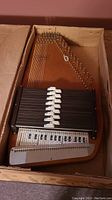 Full view of Oscar Schmidt autoharp inside original cardboard box showing the wooden body, strings, chord bars, and tuning pins.