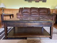 Front view of dark wood rectangular coffee table on a patterned rug, showing the lower shelf and solid legs. Sofa and side table in background.