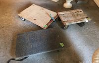 Three flat wood dollies on garage floor, two carpeted tops and one bare wood top with handle carved in. Wheels visible.