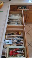 Three open wooden drawers displaying kitchen utensils, multiple flatware sets organized in plastic trays, and various hand tools.