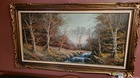 Full view of the framed oil painting showing a river flowing through a forest with mountains in the background.