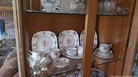 Display of the Royal Albert Petit Point china including large square plates, teacups, saucers, creamer, sugar bowl, salt and pepper shakers arranged inside a glass cabinet.