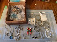 Photo of assorted jewelry including sterling silver earrings on card, colorful parrot earrings, pendants, and hoop earrings arranged on table with open cigar box containing additional pieces.