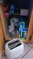 Photo of Black and Decker 2-slice toaster on floor with wooden cabinet interior showing boxes of Ziploc bags, produce bags, and other food storage items inside cabinet