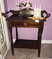 Front view of the vintage wooden wash stand showing top surface with decorative items, single drawer, lower shelf, turned legs, and casters.