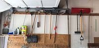 Wide shot showing various gardening and yard tools hanging on wooden board in garage wall, including multiple shovels, brooms, garden claw and rake handles.