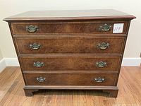 Front view of antique 4-drawer wooden dresser on wood floor with visible brass handles and drawer fronts, showing overall condition including surface scratches and scuffs.