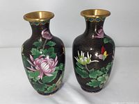 Two Chinese cloisonné vases side by side on a neutral surface, showing floral, leaf and butterfly designs on black background with gold cloud motifs and brass rims.