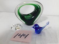 Photo showing the three glass pieces together: a large green Holmegaard art glass, a small blue art glass bird, and a clear crystal heart paperweight with a lot number card adjacent.