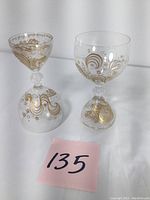 Two clear glass goblets sitting on white surface with gold swirling patterns, stars and script text around bowls and bases