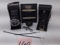 Two vintage folding cameras and associated accessories on white background. Cameras open with visible bellows and metal shutters. Accessories laid out including cables and a lens.