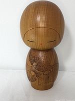 Front view of the wooden Kokeshi doll showing minimal painted facial features and engraved leaf-like decorations on the body.